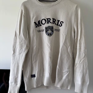 Beige stickad tröja från Morris - Snygg beige stickad tröja från Morris med broderad logga och emblem på bröstet. Tröjan har rund halsringning, långa ärmar och ribbade muddar vid ärmslut och nederkant. Perfekt för dig som gillar klassisk och stilren look.