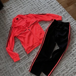 Svarta träningsbyxor med neonrosa zip-up - Snygga svarta träningsbyxor med neonrosa ränder längs sidorna. Byxorna har elastisk midja och är gjorda i ett glansigt syntetmaterial som känns sportigt och bekvämt. Tillsammans med neonrosa zip-up. Perfekta för dig som gillar en färgstark och sportig stil.