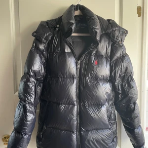 Svart dunjacka från Polo Ralph Lauren - Snygg svart dunjacka från Polo Ralph Lauren med glansig finish, röd broderad logga på bröstet och dragkedja framtill. Jackan har hög krage, avtagbar huva och två fickor med dragkedja. Perfekt för kalla dagar när du vill hålla stilen.