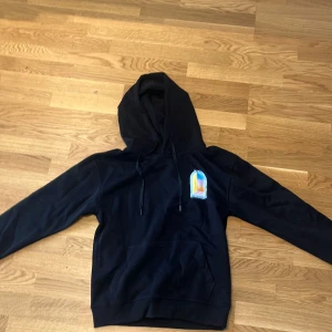 Svart hoodie från Casablanca Tennis Club - Svart hoodie från Casablanca Tennis Club med stor färgglad grafik på ryggen och mindre tryck på bröstet. Tröjan har huva med snörning, känguruficka och långa ärmar. Materialet är mjuk bomull och passformen är normal. Perfekt för dig som gillar streetwear och snygga prints.