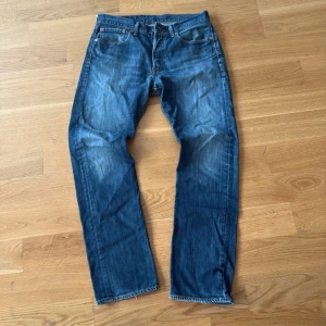Vintage Levi's 501  - Klassiska Levi's 501 jeans i blå tvätt med snyggt slitna detaljer och raka ben. Modellen har fem fickor, knappgylf och den ikoniska läderpatchen bak i midjan. Jeansen är tillverkade i robust denim och har en tidlös straight passform.