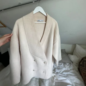 Beige kofta från Zara med knappar - Snygg beige kofta från Zara med bred ribbad krage och dubbelknäppning framtill. Koftan har en loose passform och är tillverkad i ett mjukt, stickat material som känns lyxigt och passar perfekt till lager-på-lager. Långärmad och stilren design. Brukar även ha som jacka 