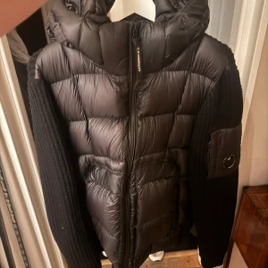 Svart pufferjacka från C.P. Company - Säljer en svart pufferjacka från C.P. Company med huva och stickade ärmar i merinoull. Jackan har quiltad front, dragkedja och en ikonisk linsdetalj på ärmen. Perfekt för dig som vill ha en snygg och modern look med både värme och stil.