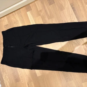 Svarta kostymbyxor med rak passform - Snygga svarta kostymbyxor med klassisk rak passform och pressveck. Byxorna har hög midja, diskreta fickor och stängs med knapp och dragkedja. Perfekta för en clean och stilren look.