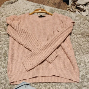 Ljusrosa stickad tröja med volang - Supersöt ljusrosa stickad tröja från Jennyfer i storlek XS. Tröjan har långa ärmar och snygga volangdetaljer på axlarna som ger en trendig touch. Perfekt för dig som gillar en feminin och mysig stil.