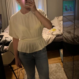 Ljusgul blus med volang och resår - Säljer en söt ljusgul blus med korta ärmar och volang nedtill. Blusen har resår i midjan som ger en snygg siluett och är tillverkad i ett mjukt tyg. Perfekt att styla med jeans eller kjol för en fräsch look.