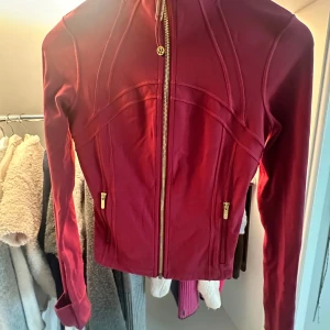 Vinröd träningsjacka från Lululemon - Säljer en vinröd träningsjacka från Lululemon med snygga sömdetaljer och guldiga dragkedjor. Jackan är figursydd, har hög krage och långa ärmar. Materialet är en mix av nylon, elastan och polyester vilket gör den stretchig och bekväm. Perfekt för aktiva dagar! Storlek 4!! knappast använd!!