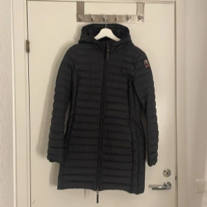 Svart dunjacka från Parajumper Irene - Svart lång dunjacka från Parajumpers med huva och snyggt emblem på ärmen. Jackan har fyra fickor på utsidan och en inneficka. Jackan är superlätt, har dragkedja framtill och två fickor med dragkedja. Perfekt för kalla dagar när du vill hålla stilen och ändå vara varm.
