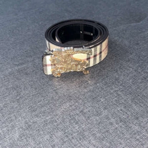 Rutig Burberry-bälte med guldfärgat spänne - Snyggt bälte från Burberry med klassiskt rutigt mönster i beige, svart, vitt och rött. Bältet har ett detaljerat guldfärgat spänne med Burberrys ikoniska riddare. Perfekt accessoar för att lyfta din outfit med en lyxig touch.