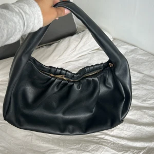 Svart axelväska i skinnimitation från pieses - Snygg svart axelväska med mjuk, rynkad design och guldfärgad dragkedja. Väskan har ett brett handtag som gör den enkel att bära över axeln och är tillverkad i skinnimitation. Perfekt för att lyfta din outfit med en cool vibe.