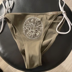 Beige bikiniunderdel från Skiny strl 38 - Snygg beige bikiniunderdel från Skiny i storlek 38 med vita knytband i sidorna. Framsidan har ett coolt tryck med texten 'Soul in your self' och grafiskt motiv. Materialet är mjukt och stretchigt, perfekt för strandhäng. Passar dig som gillar enkel men unik stil.