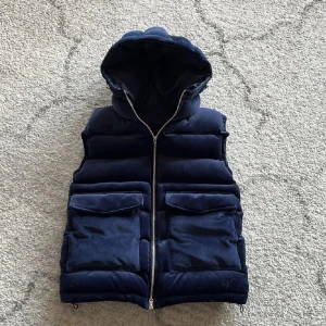 Mörkblå manchester vest med huva -  HELT NY Snygg mörkblå dunväst i manchester. Västen har en praktisk dragkedja och stora fickor framtill. Perfekt för kyliga dagar med sin quiltade design och huva.Som sagt västen är helt ny och är bara testad. Den skulle nog passa nån som är cirka 175cm vid fler frågor är det bara slå en signal🙌👑