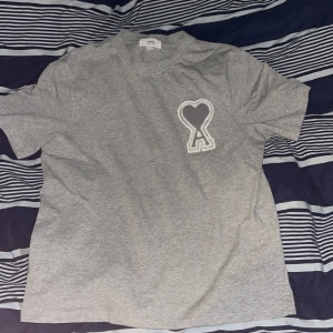 Grå t-shirt från Ami med hjärtdetalj - Säljer en grå t-shirt från Ami med klassisk rund hals och korta ärmar. På bröstet sitter en stor broderad patch med ett hjärta och bokstaven A. T-shirten är gjord i mjuk bomull och har en clean, chill vibe som funkar till jeans eller shorts. Aldrig använd 