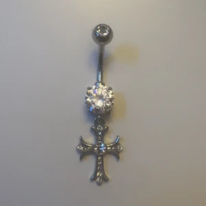 Navelpiercing med kors och strass - Snygg navelpiercing i silverfärgad metall med ett glittrigt kors hängande och stora strasstenar. Korset har flera små stenar som ger extra bling och cool detalj. Perfekt för dig som vill ha något som sticker ut och glänser.