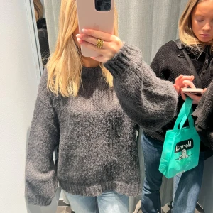Mörkgrå stickad tröja med ballongärm - Mysig mörkgrå stickad tröja med breda ballongärmar och rund halsringning. Tröjan har en loose fit och ribbade muddar vid ärmslut och nederkant. Perfekt för lager-på-lager och chill dagar.