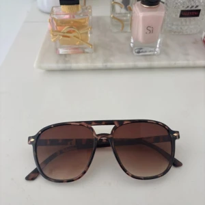 Bruna pilotbågar solglasögon leopardmönster - Snygga solglasögon med pilotformade bågar i brunt leopardmönster och bruna tonade glas. Ramen är i plast och har en cool detalj med dubbelbrygga. Perfekta för att lyfta din stil med en trendig touch.