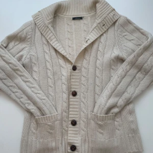 Beige kabelstickad kofta från Adam - Snygg beige kabelstickad kofta från Adam med stora bruna knappar och två fickor framtill. Koftan har bred krage och långa ärmar, perfekt för lager-på-lager. Den stickade strukturen ger en klassisk och mysig vibe.
