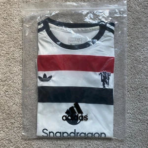 Manchester United Adidas fotbollströja - Snygg Manchester United fotbollströja från Adidas med breda ränder i rött och svart på vit botten. Tröjan har korta ärmar, rund hals och svarta detaljer på axlarna. Materialet är lätt och andas, perfekt för träning eller match. Loggor från Adidas och Snapdragon på framsidan.