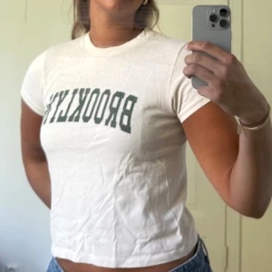 Beige t-shirt - Beige t-shirt med grön BROOKLYN-tryck på bröstet från Pure Cashmere. Modellen är kortärmad och har en klassisk rund hals. T-shirten är gjord i mjuk bomull och har en normal passform som sitter snyggt till jeans eller shorts.