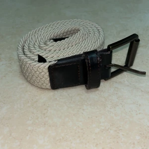 Beige flätat bälte med skinn-detaljer - Snyggt beige flätat bälte med mörkbruna skinn-detaljer vid spännet. Bältet har ett klassiskt metallspänne i matt finish och passar perfekt till jeans eller chinos. Den flätade designen ger en modern och stilren look.