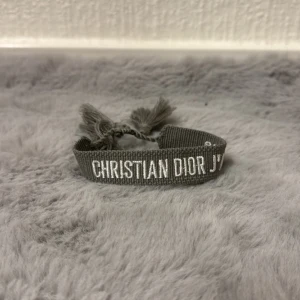 Grå Christian Dior J'Adior armband - Snyggt grått armband från Christian Dior med vit broderad text 'CHRISTIAN DIOR J'ADIOR'. Armbandet är gjort i ett mjukt tygmaterial och har tofsar i ändarna för en cool och avslappnad look. Perfekt accessoar för att lyfta din outfit.