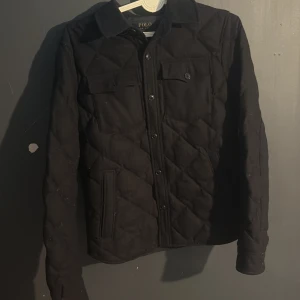 Svart quiltad jacka Polo Ralph Lauren - Quiltad svart jacka från Polo Ralph Lauren i klassisk skjortmodell. Jackan har knappar framtill, två bröstfickor med lock och knapp samt sidofickor. Snyggt rutmönster i quiltningen och mjukt material som känns lyxigt. Perfekt för dig som gillar stilren och clean look.