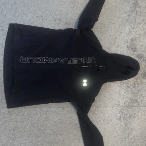 Svart vindjacka från Under Armour - Svart vindjacka från Under Armour med huva och reflekterande detaljer. Jackan har dragkedja framtill, stora logotyper och är tillverkad i lätt syntetmaterial. Perfekt för löpning eller utomhusaktiviteter när det blåser. Snygg och sportig look!