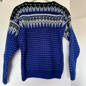 Handgjord stickad tröja - Säljer en handgjord blå stickad tröja med vitt och svart mönster över axlar och bröst. Perfekt för kyliga dagar och har en klassisk nordisk vibe med sitt grafiska mönster. Använd endast en gång