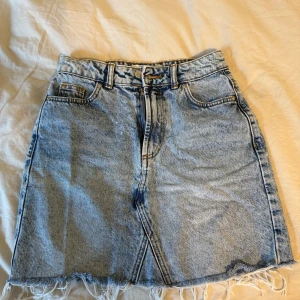 Ljusblå jeanskjol med fransig kant - Säljer en snygg ljusblå jeanskjol med hög midja och fransig nederkant. Kjolen har klassiska fickor fram och bak samt bälteshällor. Perfekt för dig som gillar en avslappnad och trendig stil.