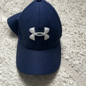 Blå keps från Under Armour - Snygg marinblå keps från Under Armour med klassisk böjd skärm och stort vitt broderat logotyp framtill. UA Classic Fit-modell med ventilerande material och bekväm passform. Perfekt för sport eller chill dagar utomhus.