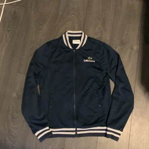 Marinblå bomberjacka från Lacoste Sport - Snygg marinblå bomberjacka från Lacoste Sport med vita ränder på krage, ärmslut och midja. Jackan har dragkedja framtill, två fickor och klassisk Lacoste-logga på bröstet. Tillverkad i polyester och bomull för en sportig och clean look.