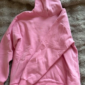 Rosa hoodie med huva - Säljer en enkel och snygg rosa hoodie med huva. Tröjan har långa ärmar, ribbade muddar och är gjord i mjukt material som känns skönt mot huden. Perfekt för dig som gillar en chill och avslappnad stil.