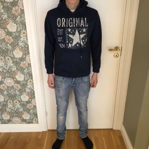 Mörkblå hoodie från original - Snygg mörkblå hoodie från Original Fit med stort vitt tryck och stjärndetalj på bröstet. Tröjan har huva med snörning och en klassisk känguruficka framtill. Perfekt för dig som gillar en avslappnad och cool stil.