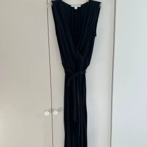 Svart omlott jumpsuit med knyt - Svart jumpsuit från DVF med omlott-v-ringning och knyt i midjan. Klänningen är ärmlös och har ett mjukt, fallande tyg som ger en snygg siluett. Perfekt för dig som gillar stilrena och eleganta plagg med lite edge. Jumpsuiten är i storlek US 8