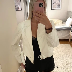 Vit croppad skinnjacka med ringdetalj - Snygg croppad vit skinnjacka från Bershka med cool ringdetalj vid dragkedjan och knappar vid midjan. Jackan har långa ärmar, markerad krage och stilren design som ger en edgy vibe till din outfit.