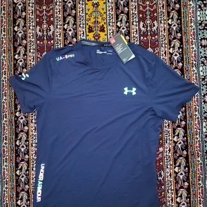 Blå Under Armour HeatGear t-shirt - Snygg marinblå t-shirt från Under Armour med UA-logga på bröstet och tryck på axeln. Modellen är kortärmad och har rund hals. Materialet är lätt och andas, perfekt för träning. Fitted passform som sitter skönt mot kroppen. UA HeatGear-teknologi för extra komfort.