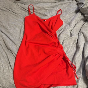 Röd omlottklänning från Divided - Snygg röd kortklänning från Divided med omlott-design och smala axelband. Klänningen har en cool snörningsdetalj på sidan som ger en trendig look. Perfekt för dig som vill sticka ut med färg och stil. Materialet känns mjukt och lätt mot huden.