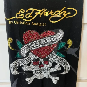 Ed Hardy svart klänning med dödskalle - Svart Ed Hardy klänning med ikoniskt dödskalle- och hjärtmotiv framtill, glittriga detaljer och guldtext. Ärmlös modell med brottarrygg och tryck även på baksidan. Perfekt för dig som gillar edgy och unik streetstyle.