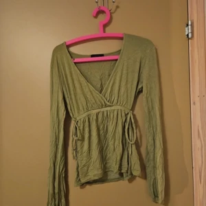 Olivgrön topp från Vero Moda - Olivgrön långärmad omlottopp från Vero Moda med knytband i midjan. Djup v-ringning och mjukt, lätt material som ger en avslappnad men feminin siluett. Köpt på Vinted, men jag har bara använt den 1 gång. Storlek M.