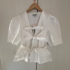Vit blus med rosetter från Ganni - Helt ny, supersöt vit blus från Ganni med tre stora rosetter framtill och kort puffärm. Blusen har v-ringning och är super somrig. Den är storlek 42 men passar mig som oftast har storlek S/XS perfekt då man kan knyta den ❣️Nypris 2599 💗
