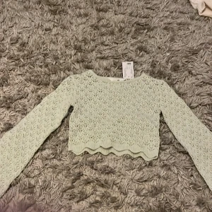 Ljusgrön croppad stickad tröja  - Säljer en ljusgrön croppad stickad tröja från H&M. Tröjan har lång ärm, vågig nederkant och ett fint spetsliknande mönster. Perfekt för dig som gillar en söt och trendig stil!