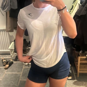 Vit t-shirt från Gymshark - Säljer en klassisk vit t-shirt från Gymshark med rund hals och diskret logga på bröstet. Perfekt för träning eller en sportig look. T-shirten är i mjuk bomull och har en normal passform med korta ärmar. Slits på sidan som går att knyta eller bara ha som den är. Säljer likadan i svart☺️