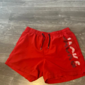Röda badshorts från Jack & Jones - Snygga röda badshorts från Jack & Jones med svartvit snörning i midjan och stort tryck längs benet. Klassisk passform med elastisk midja och innerbyxa i mesh. 