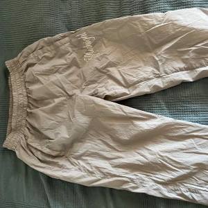 Beige mjukisbyxor från Active Boys - Beiga mjukisbyxor från Active Boys med resår i midjan och vit broderad text på ena benet. Byxorna har sidofickor och är tillverkade i ett lätt syntetmaterial med meshfoder för extra komfort.