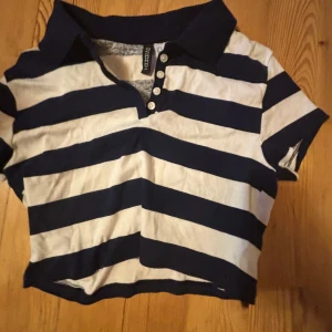 Randig croppad pikétröja från H&M Divided - Säljer en croppad pikétröja från H&M Divided i marinblått och vitt. Tröjan har breda horisontella ränder, korta ärmar, krage och knappar framtill. Perfekt för en sportig och avslappnad stil.