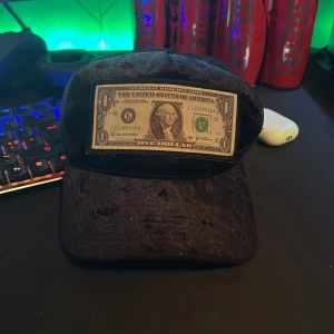 Svart keps med dollartryck - Unik svart keps med en fastsydd amerikansk dollarsedel framtill. Kepsen har klassisk böjd skärm och justerbar baksida i mesh-material för bra ventilation. Perfekt för dig som vill sticka ut med en annorlunda accessoar.