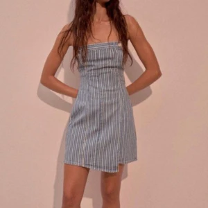Blåvit randig axelbandslös klänning - Säljer en blåvit axelbandslös kortklänning med smala vertikala ränder. Klänningen har en rak, tight passform och en liten slits framtill. Perfekt för varma dagar och enkel att styla med sneakers eller sandaler.