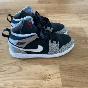 Nike Air Jordans i svart och grått - Säljer ett par Nike Air Jordans i svart och grått med vit sula och vit swoosh. Skorna har svarta skosnören, grått mönster på tån och hälen samt röd Jumpman-logga på plösen. Perfekta för dig som gillar streetwear och sneakers.
