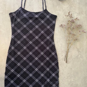 Svart rutig kortklänning från H&M - Säljer en svart kortklänning från Divided med smala axelband och vitt rutmönster. Klänningen har en rak passform och är tillverkad i mjuk polyester. Perfekt för dig som gillar en enkel men stilren look.