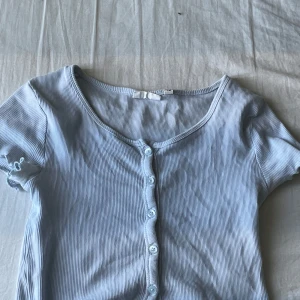 Ljusblå  croppad topp med knappar - Säljer en ljusblå croppad topp med korta ärmar och knappar framtill. Toppen är ribbad och har volangdetaljer vid ärmslut. Tillverkad i bomull och elastan för en mjuk och stretchig känsla. ❤️